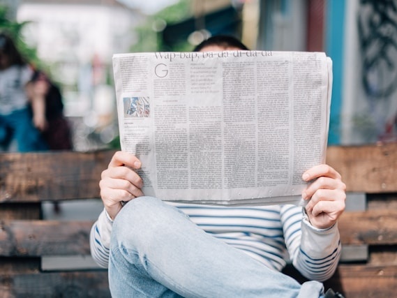 Ein Mensch sitzt auf einer Holzbank und hält eine aufgeschlagene Zeitung vor das Gesicht, sodass es nicht sichtbar ist. Die Person trägt ein gestreiftes Oberteil und Jeans. Im Hintergrund sind bunte Graffitis und ein paar verschwommene Personen zu sehen.