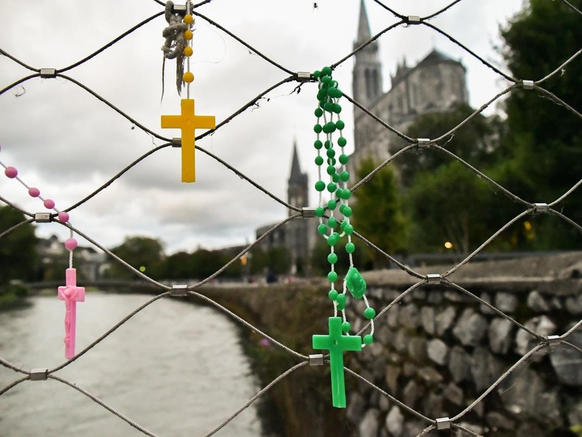 Das Bild zeigt drei Rosenkränze, die an einem Metallzaun hängen. Ein pinker Rosenkranz mit einem rosa Kreuz, ein gelber Rosenkranz mit einem goldenen Kreuz und ein grüner Rosenkranz mit einem grünen Kreuz. Im Hintergrund ist eine Kirche und ein Fluss zu sehen.